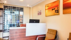 A hotel reception area with a wooden desk, a computer, a chair, and a window with a view of a building. - Grand Duke Hotel Limited (Homa Bay)