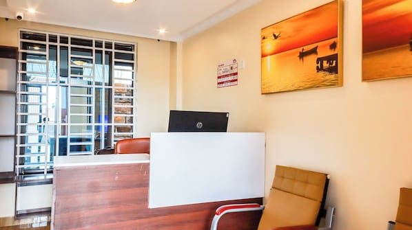 Zona de recepción de hotel con un escritorio de madera, una computadora, una silla y un ventanal con vista a un edificio.