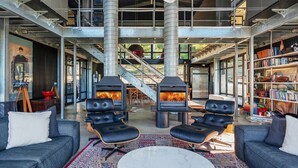 A spacious living area with a high ceiling, exposed ductwork, and a central fireplace. There are leather chairs and a coffee table, with a bookshelf filled with books and decorative items. - Sky Ridge, Kangaroo Valley (Kangaroo Valley)