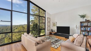 A spacious living room with large windows offering a forest view, a modern sofa set, a flat-screen TV, and a bookshelf. - Valley View House, Kangaroo Valley (Beaumont, Kangaroo Valley)