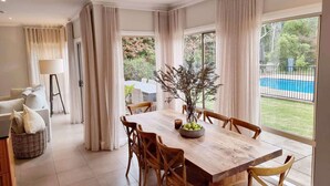 A dining area with a wooden table, chairs, and a vase with branches. There is a view of a swimming pool outside. - Westbrook, Kangaroo Valley (Kangaroo Valley)