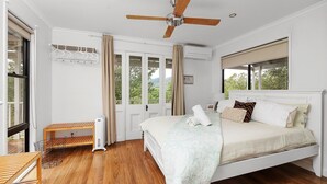 A bedroom with a bed, a ceiling fan, a window with curtains, a radiator, and a wooden bench. - Wild Rose Cottage Kiaroo Estate (Barrengarry, Kangaroo Valley)
