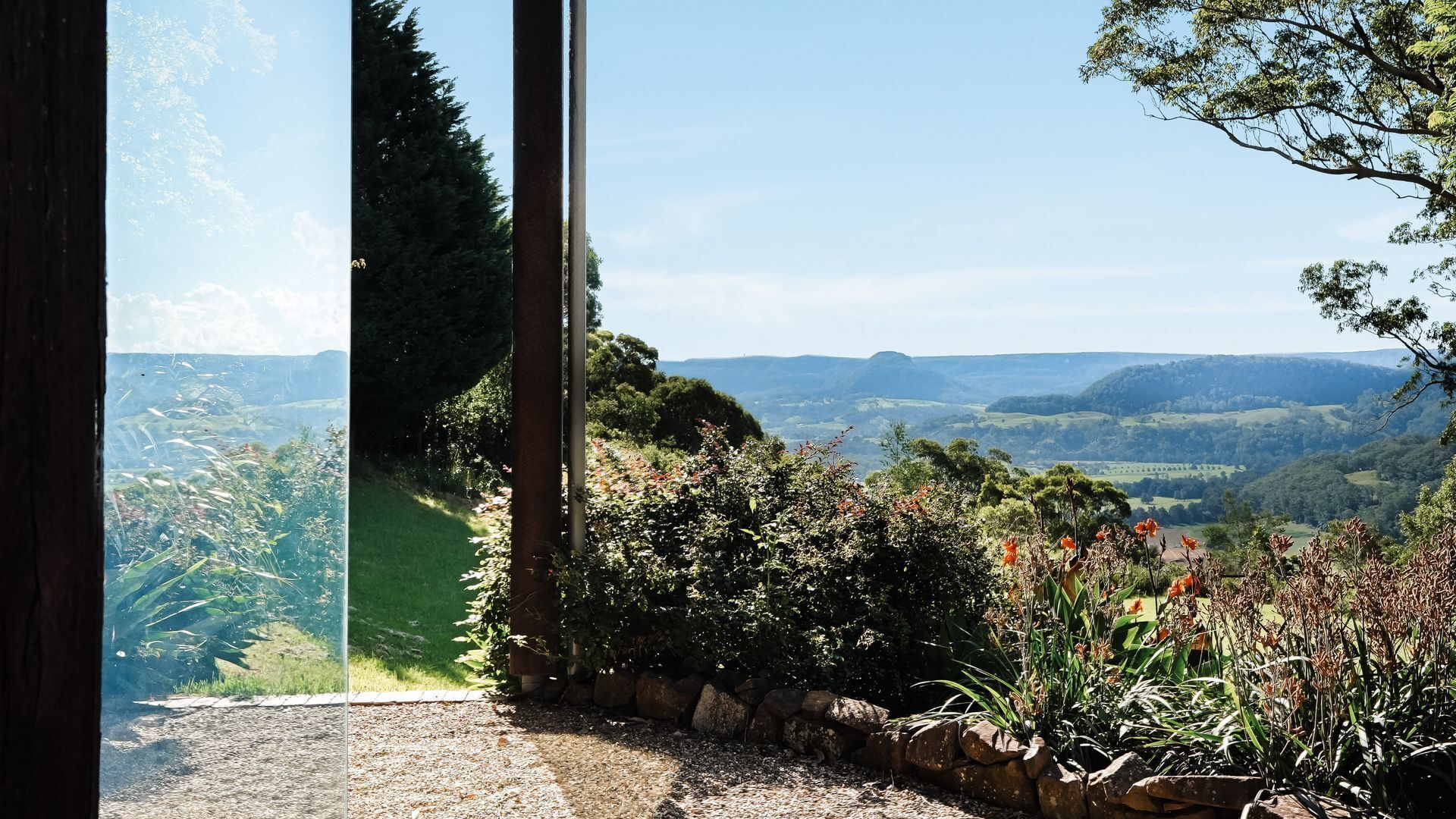 A view through a window with a stone wall and greenery, overlooking a hilly landscape.