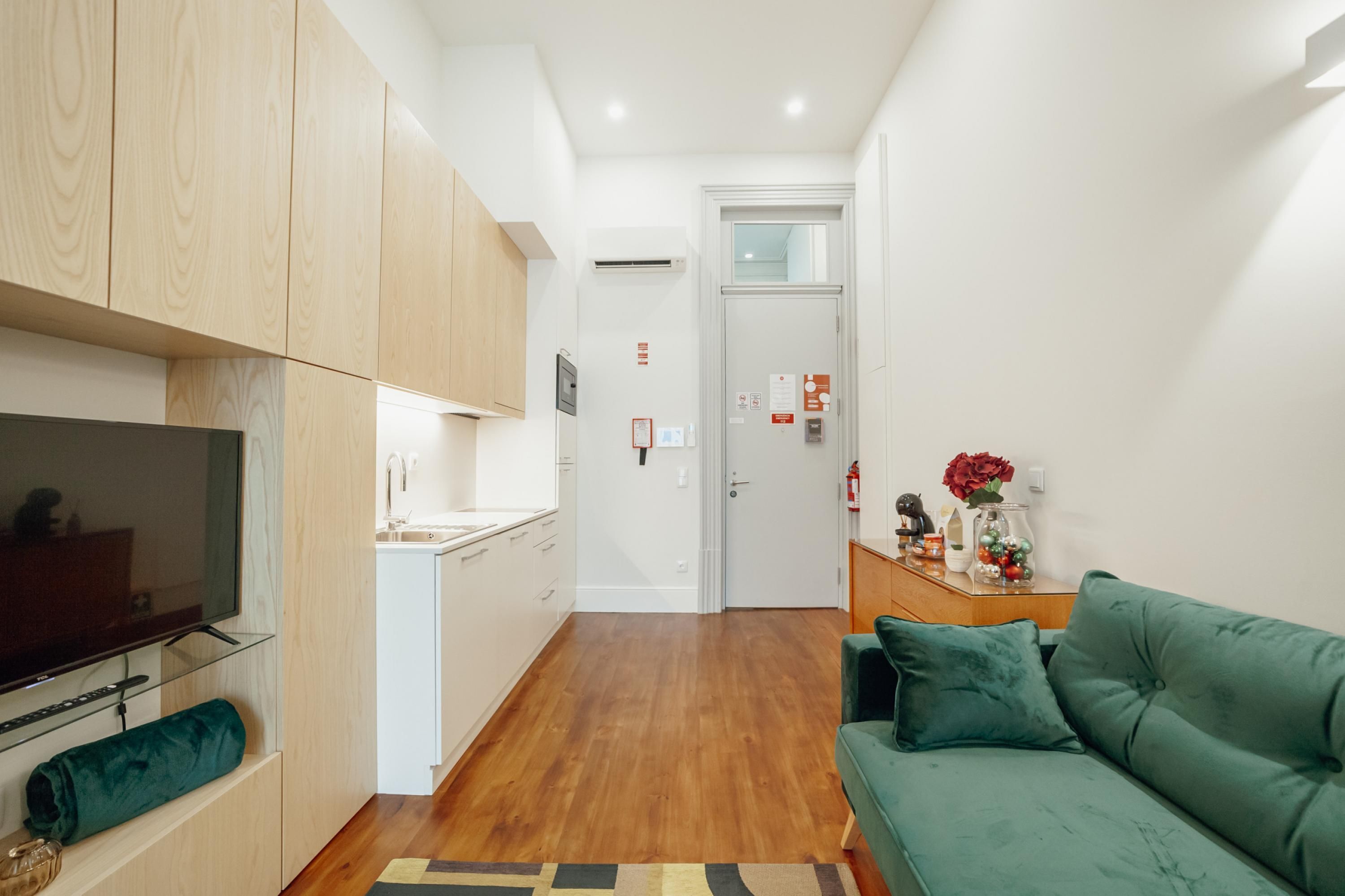 A modern living room with a green sofa, a flat-screen TV, wooden cabinets, and a kitchen area with white appliances.
