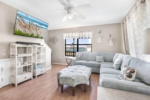 A living room with a sectional sofa, a white entertainment unit, a ceiling fan, and a large window with a view.