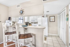 Une cuisine compacte avec des meubles blancs, un four à micro-ondes et un réfrigérateur. On y trouve deux chaises en bois avec des coussins foncés. Une horloge décorative est accrochée au mur.