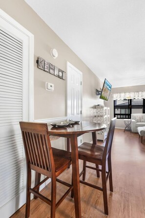 A dining area with a wooden table and chairs, a white door, and a wall-mounted television. - Heron Landing 14- Beach access, on Little Lagoon (Gulf Shores)