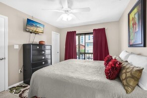 Ein Schlafzimmer mit Bett, Schminktisch, Deckenventilator, Fernseher und Fenster mit Vorhängen.