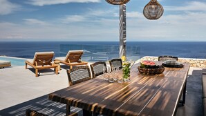 Un espace de restauration en plein air offrant une vue sur l’océan, avec des tables et des chaises en bois, des lampes suspendues en osier et un ciel dégagé.