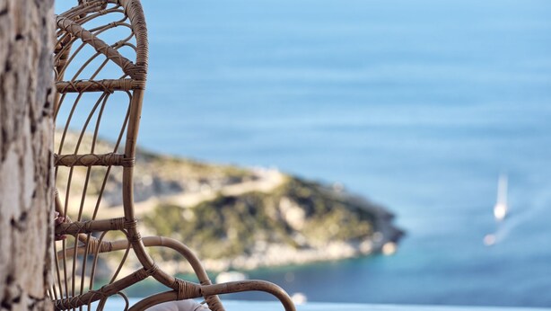 Un fauteuil suspendu en osier, doté d’un coussin, offrant une vue imprenable sur la côte, avec un ciel dégagé et une mer calme.