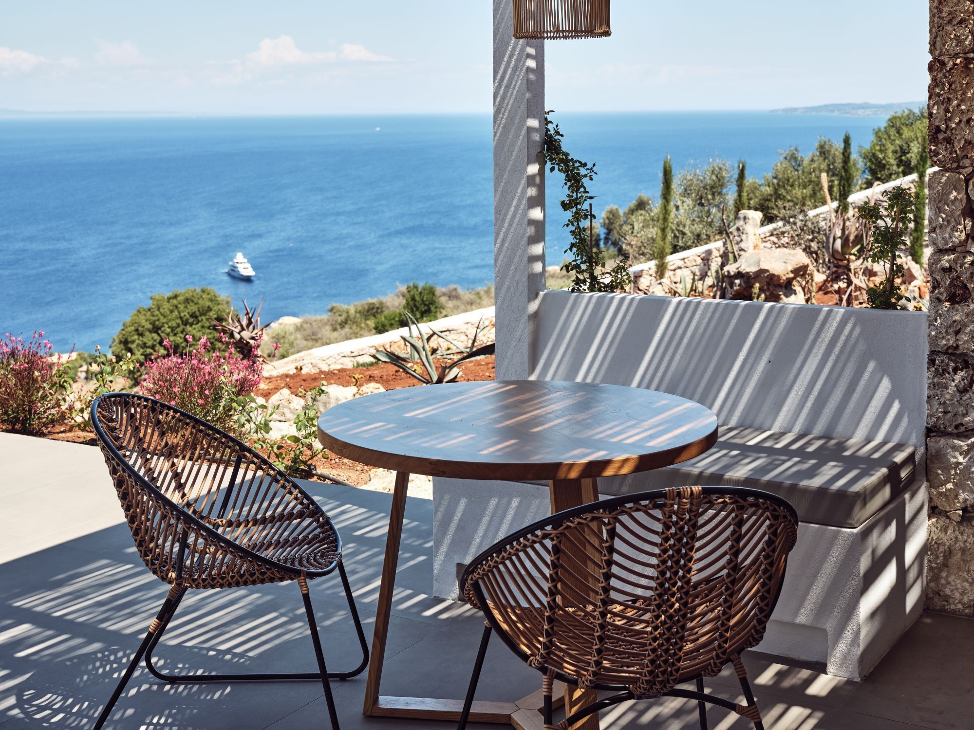En terrasse med et rundt bord og to fletstole, med udsigt over kysten med en båd i det fjerne.
