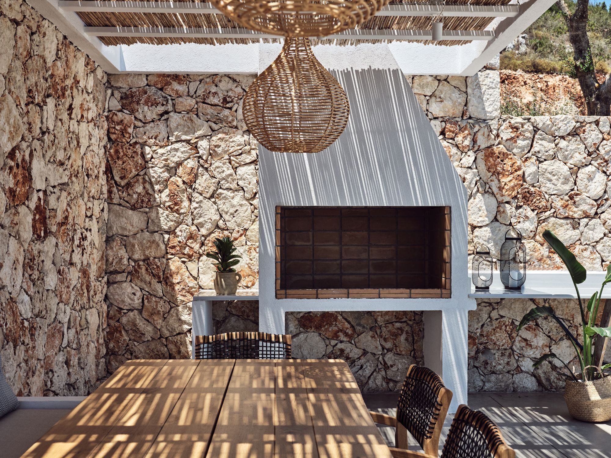 A stone wall, wooden table, wicker chairs, and a hanging light fixture in a patio area.