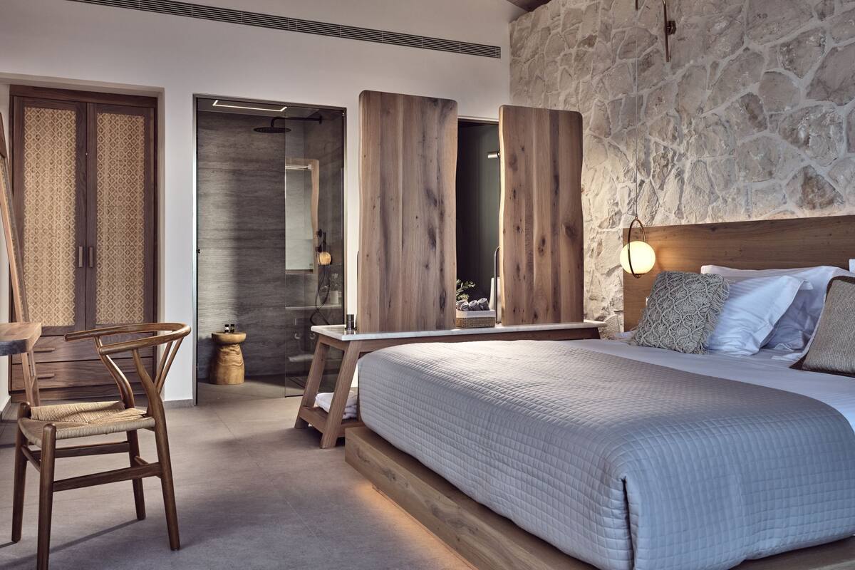 Een slaapkamer met een groot bed, houten hoofdbord, stenen muur en een klein houten tafeltje met een stoel.