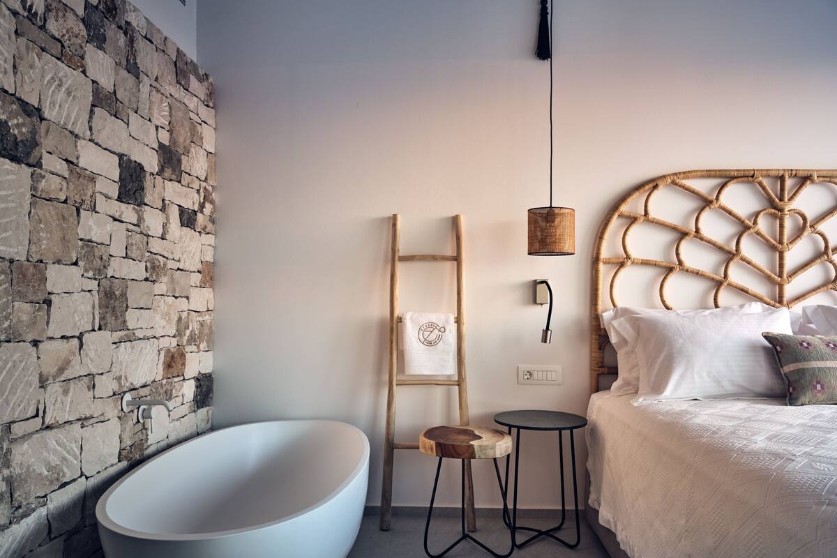 Een moderne badkamer met een stenen wand, een vrijstaand bad, een houten trapje, een klein rond tafeltje en een bed met een geweven hoofdbord.