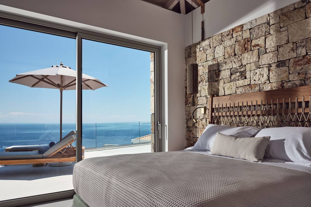 Een slaapkamer met een stenen muur, een bed met witte lakens en uitzicht op de oceaan door een glazen deur.