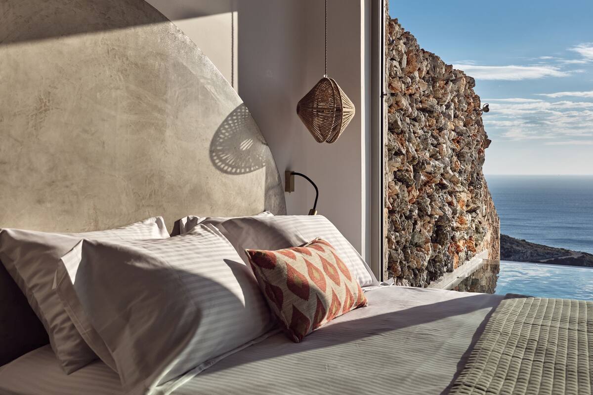 Een slaapkamer met een bed, kussens, een wandlamp en uitzicht op de oceaan.