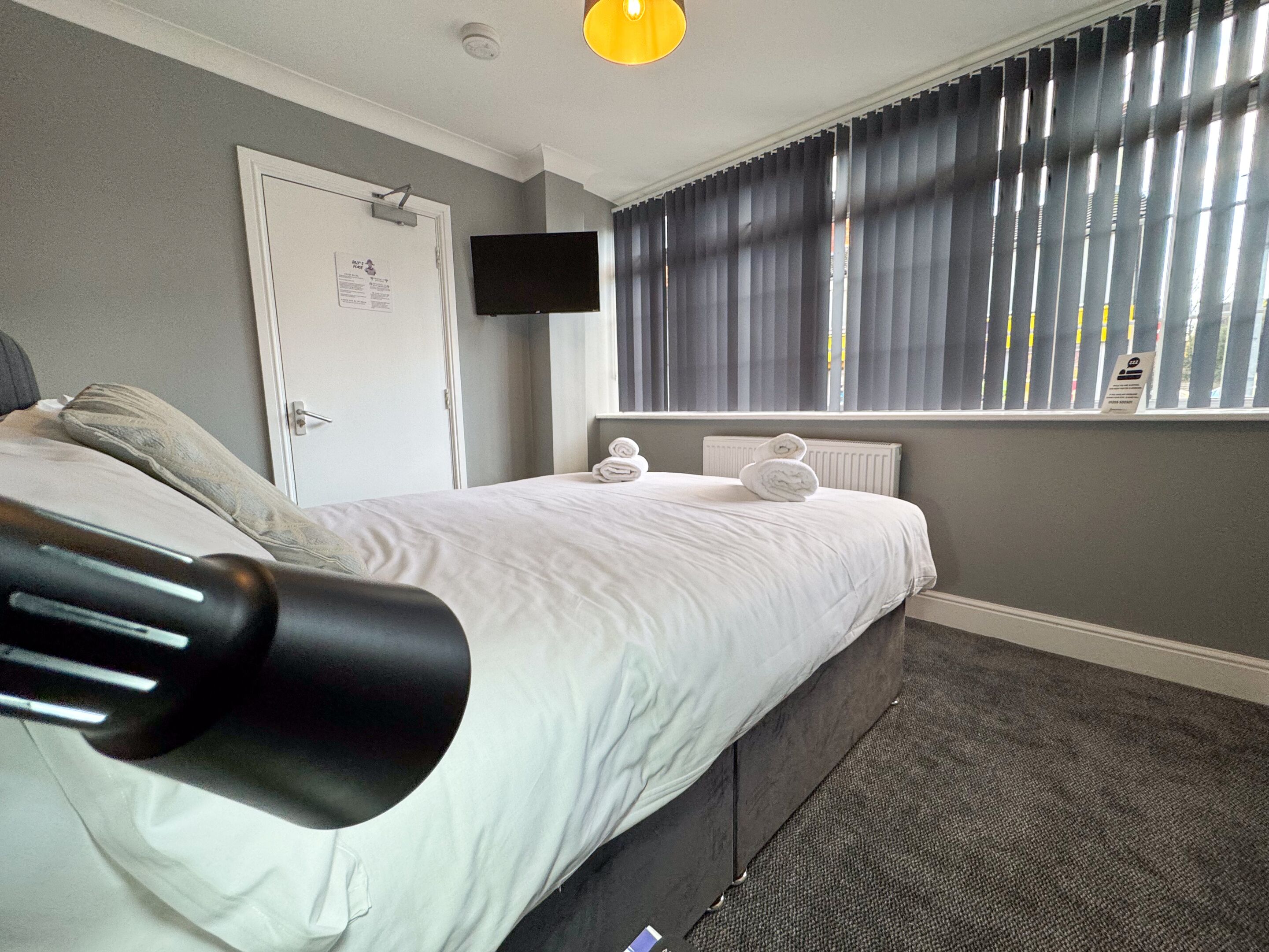 Habitación de hotel con cama, televisor de pantalla plana en la pared, ventana con persianas y radiador debajo de la ventana.