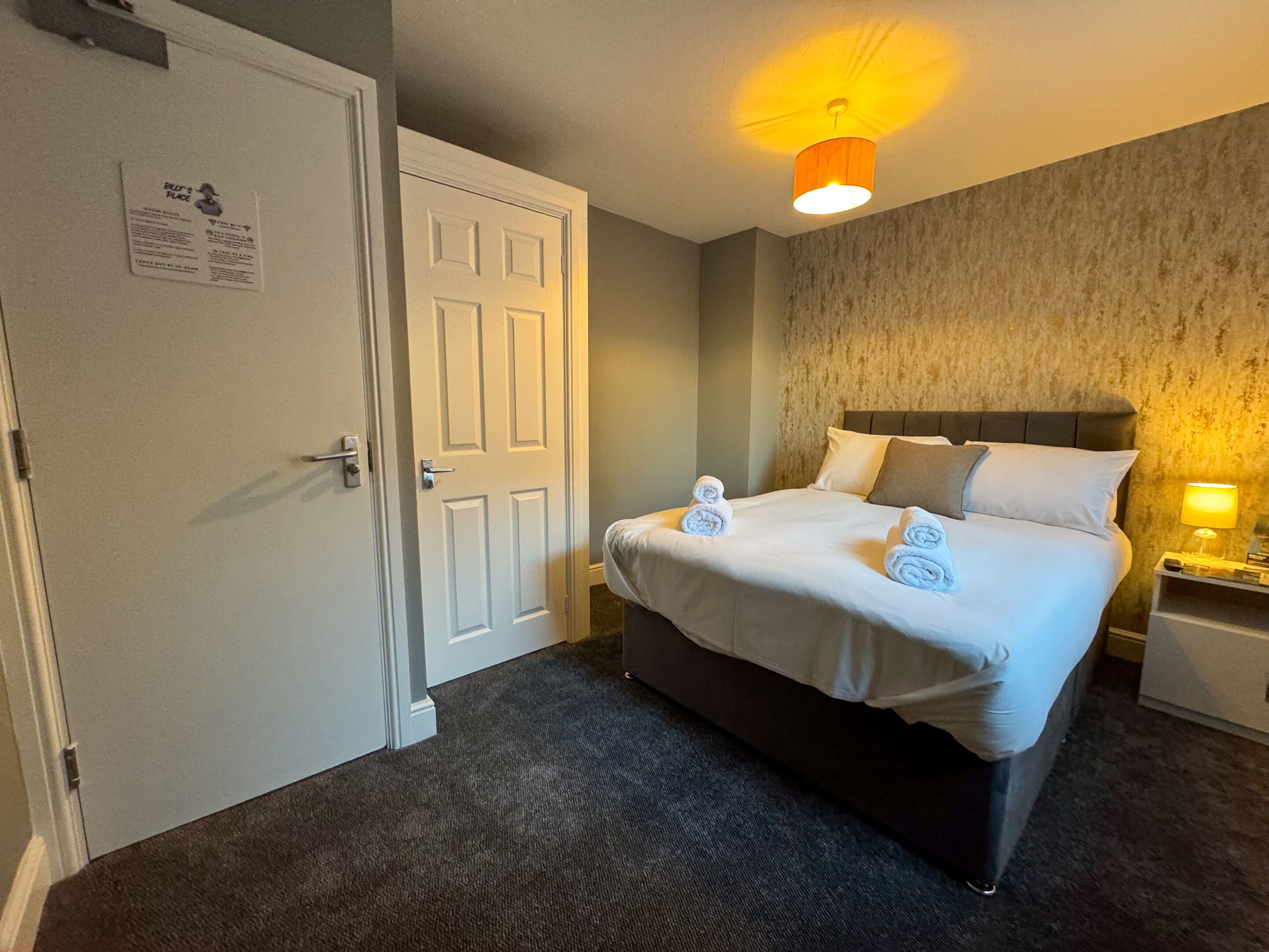 A hotel room with a bed, a nightstand, a lamp, and a door.
