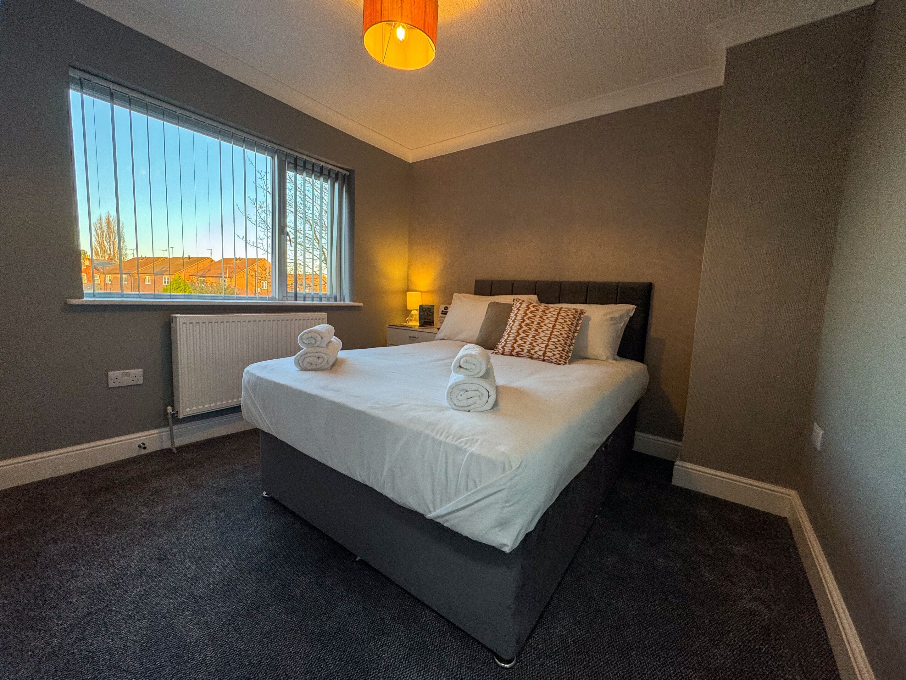 Een hotelkamer met een groot bed, een raam met jaloezieën, een radiator en een bedlamp.