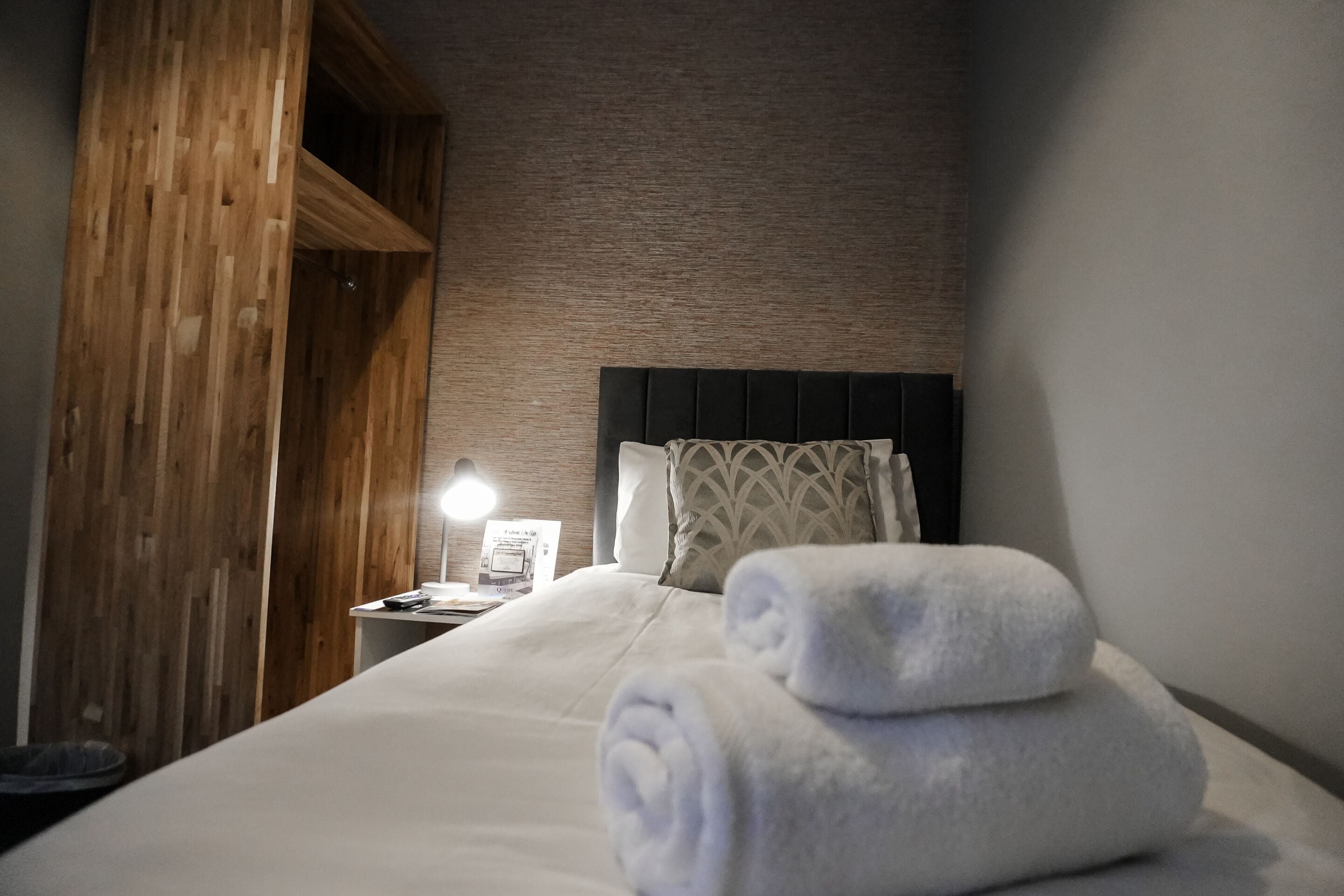 A neatly made bed with a folded towel, a bedside lamp, and a wooden wall panel.
