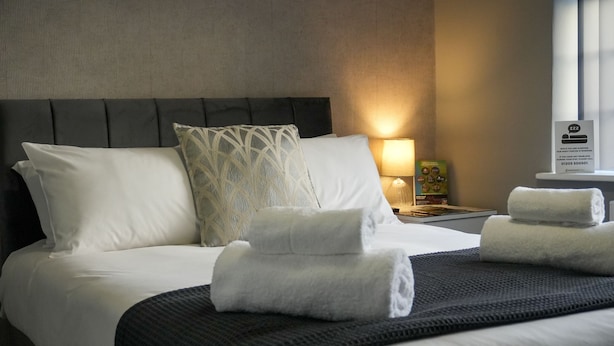 Una cama bien hecha con sábanas blancas, un cabecero oscuro y una lámpara de noche.