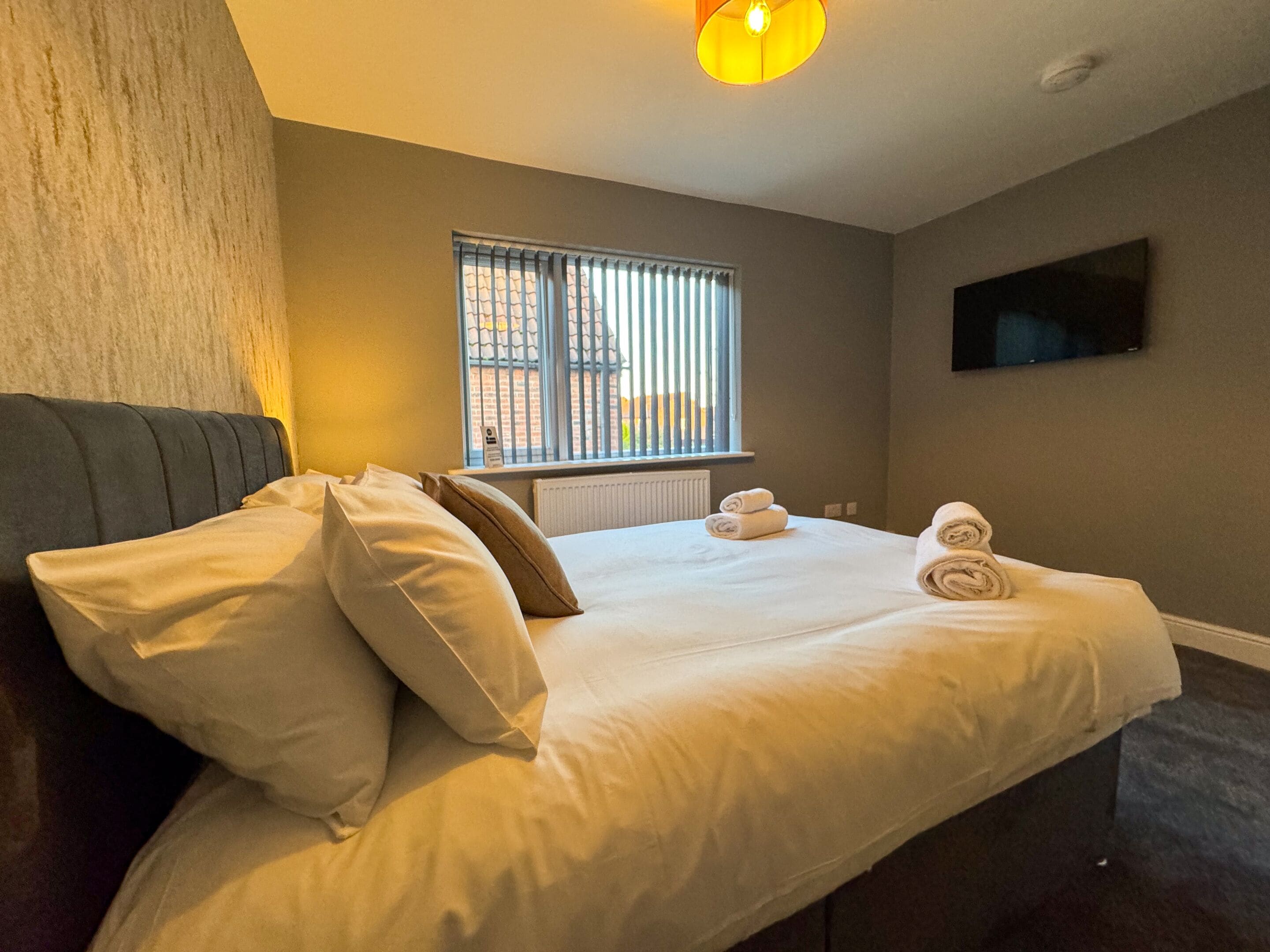 Un dormitorio con cama, almohadas, ventana con persianas, radiador y televisor de pantalla plana en la pared.
