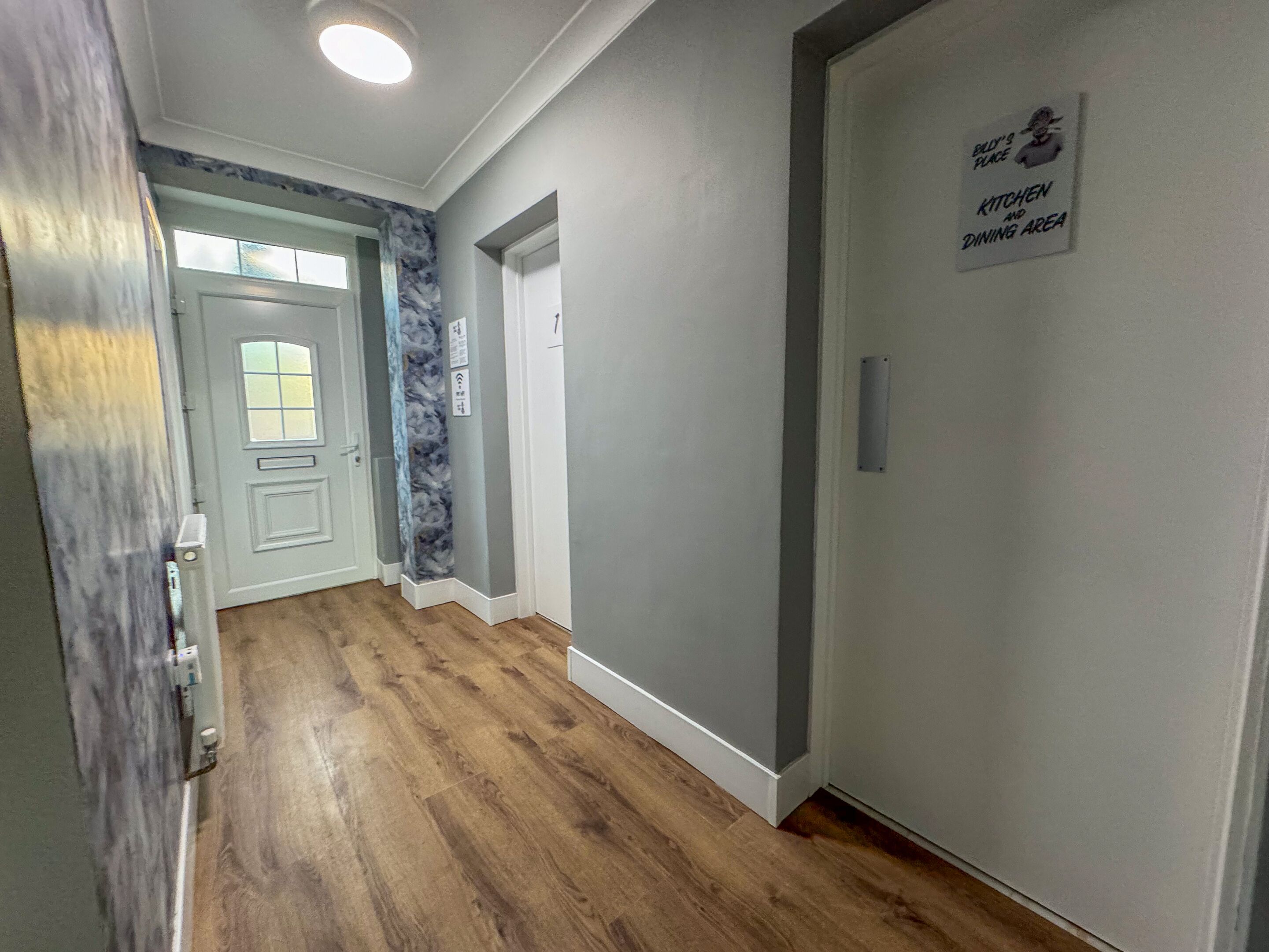A hallway with wooden flooring, a door with a frosted glass window, and a sign indicating a kitchen and dining area.
