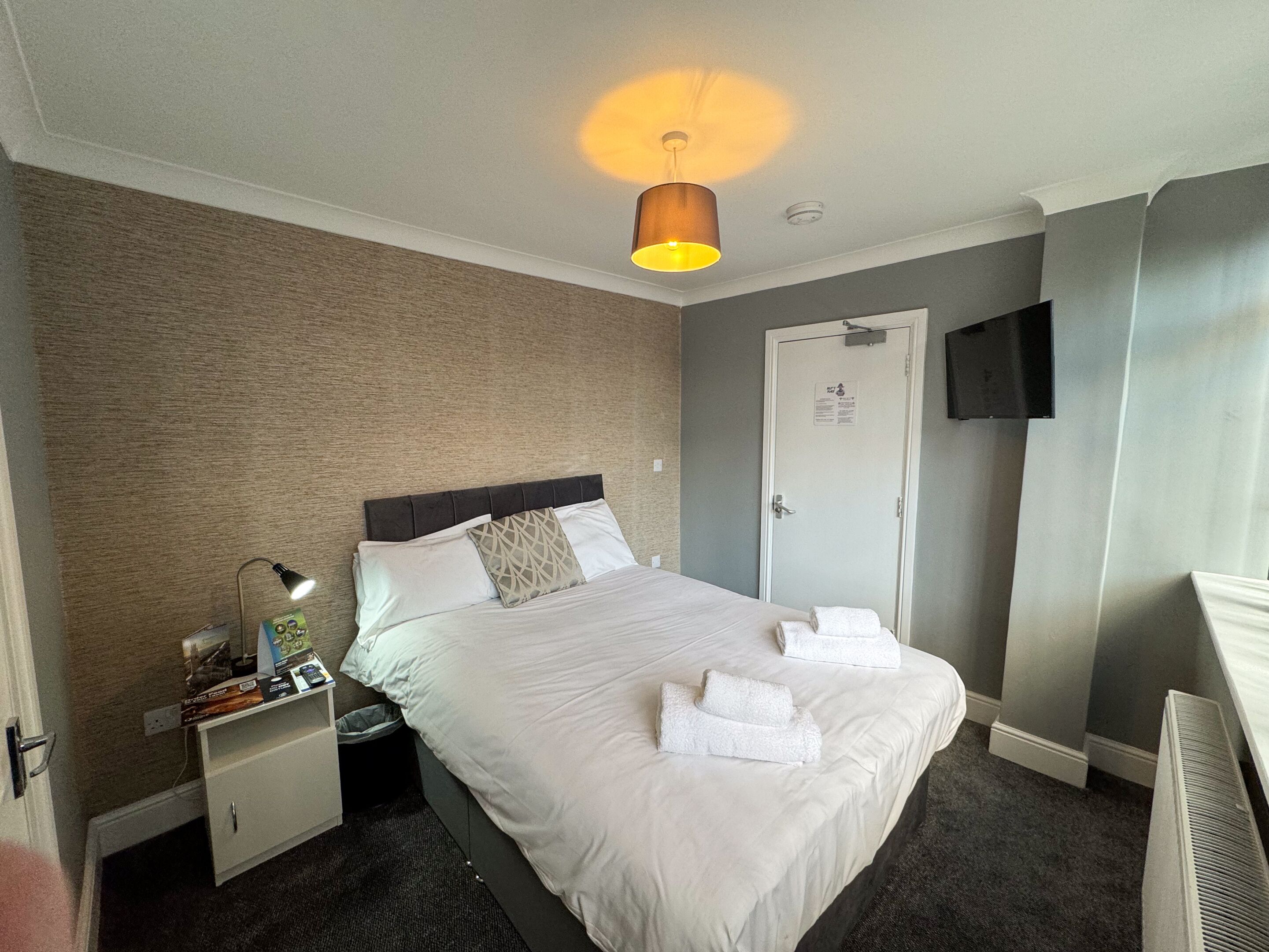 A hotel room with a bed, a nightstand, a lamp, a TV, and a door.