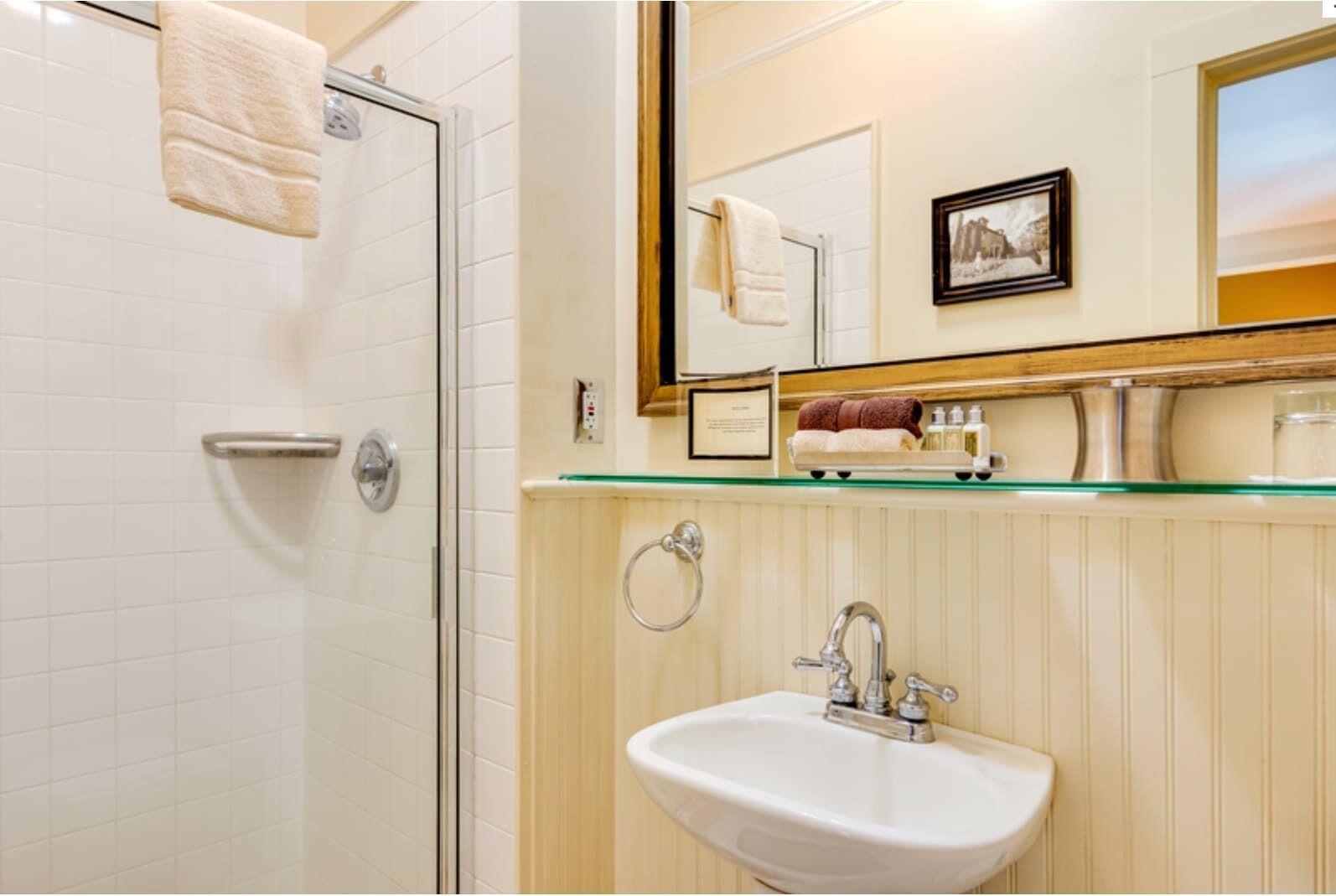 A bathroom with a glass shelf holding toiletries, a sink, and a shower area.