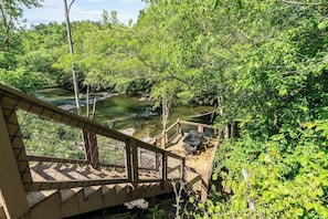 A wooden deck with a table and chairs overlooking a river, surrounded by dense greenery.