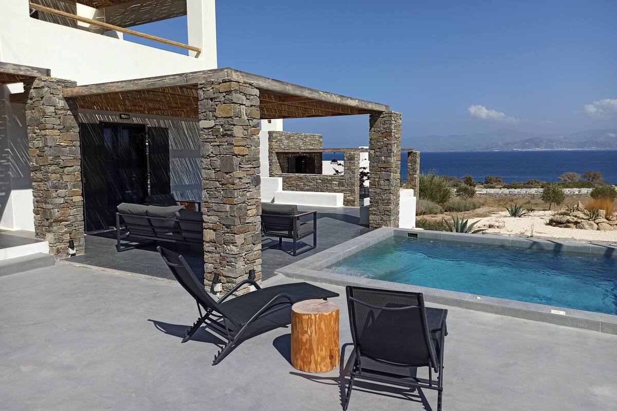 A modern outdoor pool area with stone pillars, lounge chairs, and a view of the sea.