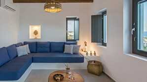 A modern living room with a blue sectional sofa, a wooden coffee table, and a view of the sea through a window. - Vastblue (Paros)