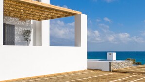A modern white building with a wooden pergola, a stone wall, and a view of the sea. - Vastblue (Paros)