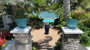 A paved pathway leading to a pool with a view of the ocean, flanked by stone pillars supporting large planters with various plants. - Saltwater Sanctuary-Secret garden in a tropical paradise (Hope Town)