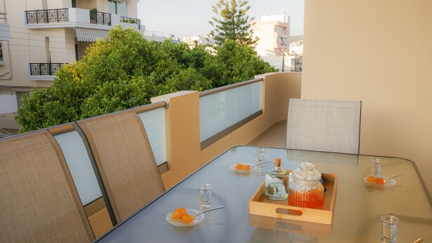 Un balcon avec une table en verre, dressée pour le dîner, sur laquelle on trouve un plateau avec des fruits oranges, un verre d’eau et une bouteille.
