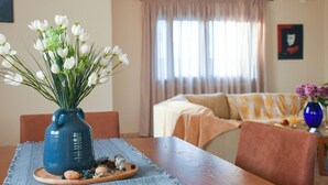 Un coin repas avec une table dressée pour deux personnes, un vase rempli de fleurs blanches et une vue sur un salon avec un canapé et des rideaux.