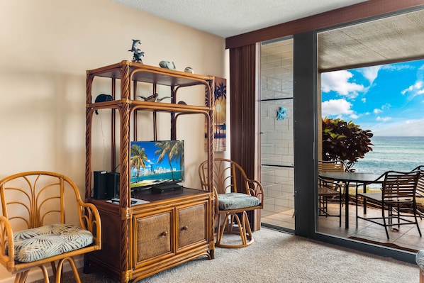 Una habitación con un mueble de televisión de mimbre, dos sillas de mimbre, una mesa de comedor con sillas y vista al océano a través de un ventanal.