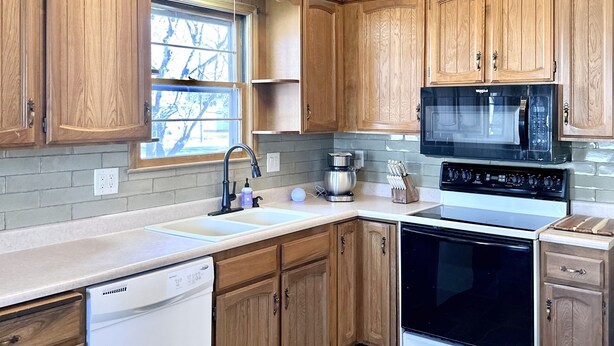 A modern kitchen with wooden cabinets, a dishwasher, an oven, a microwave, and a window with a view of trees. - Lee’s Garden House (Oskaloosa)