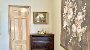 A wooden door, a dark wooden dresser with drawers, a framed painting of a house, and a floral artwork on the wall. - Lee’s Garden House (Oskaloosa)