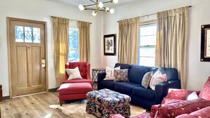 A living room with a wooden door, a red armchair, a blue sofa, and patterned ottoman. - Lee’s Garden House (Oskaloosa)