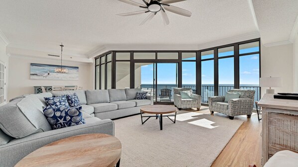 A spacious living area with a large gray sofa, patterned pillows, a wooden coffee table, and wicker chairs. A ceiling fan is visible, and the room has large windows offering a view of the ocean.