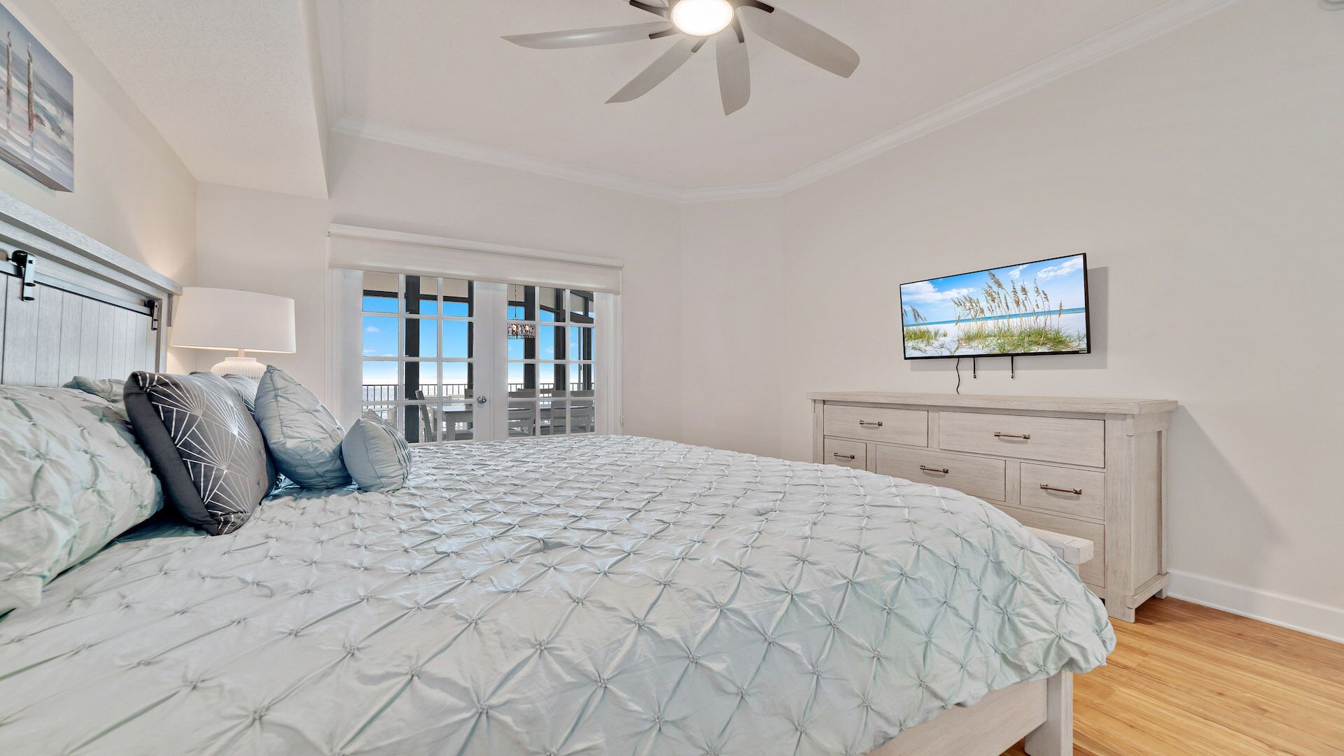 A bedroom with a bed, a TV, a ceiling fan, and a view through a glass door.