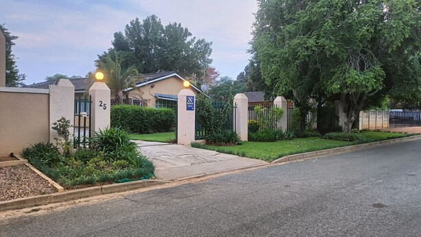 A gated residential area with a well-maintained lawn, a driveway leading to a house, and a sign indicating a National school nearby.