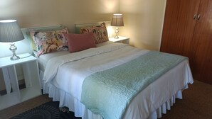 A neatly made bed with a white and light blue bedspread, two floral pillows, and a matching throw. There is a white nightstand with a lamp and a small table beside the bed. A wooden wardrobe is visible in the background.