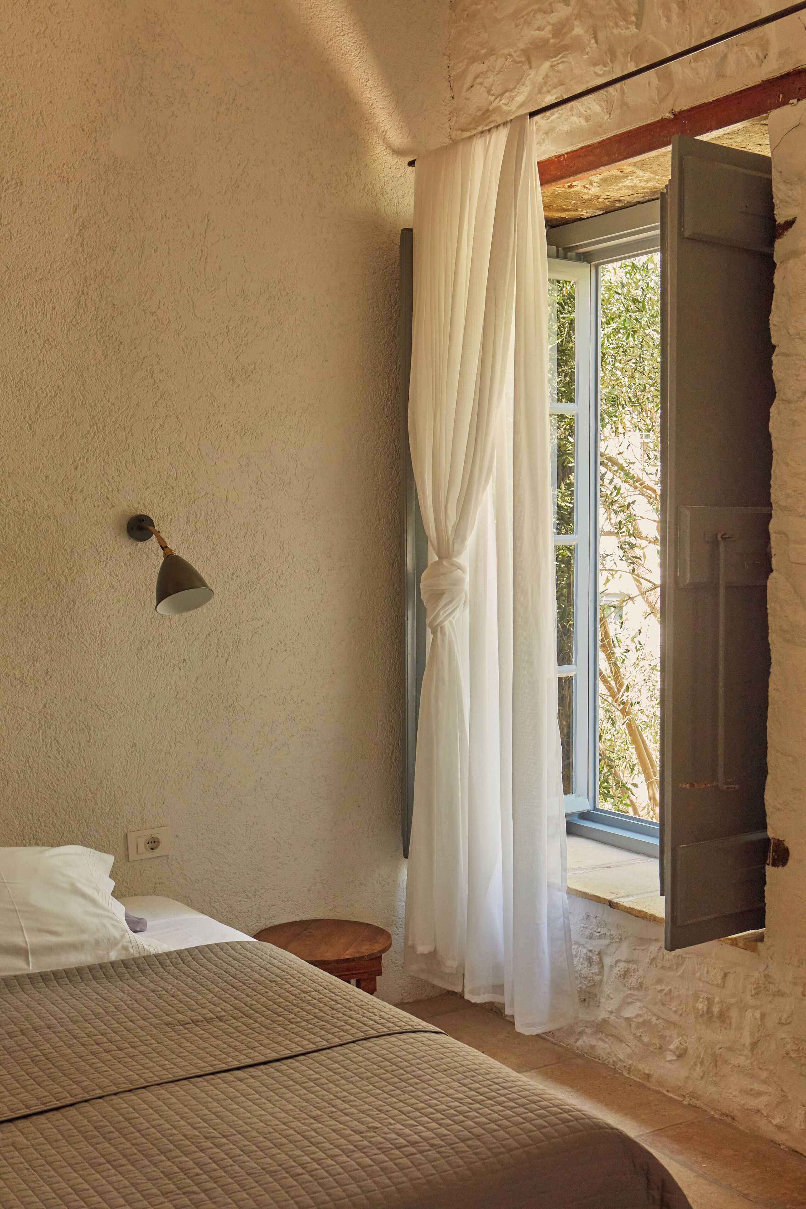 A bedroom with a bed, a bedside table, a wall-mounted lamp, a window with curtains, and a view of greenery outside.