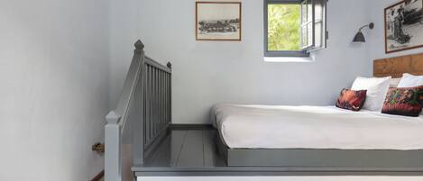 Un dormitorio con una cama, una ventana pequeña, un cuadro en la pared y una mesita de noche.