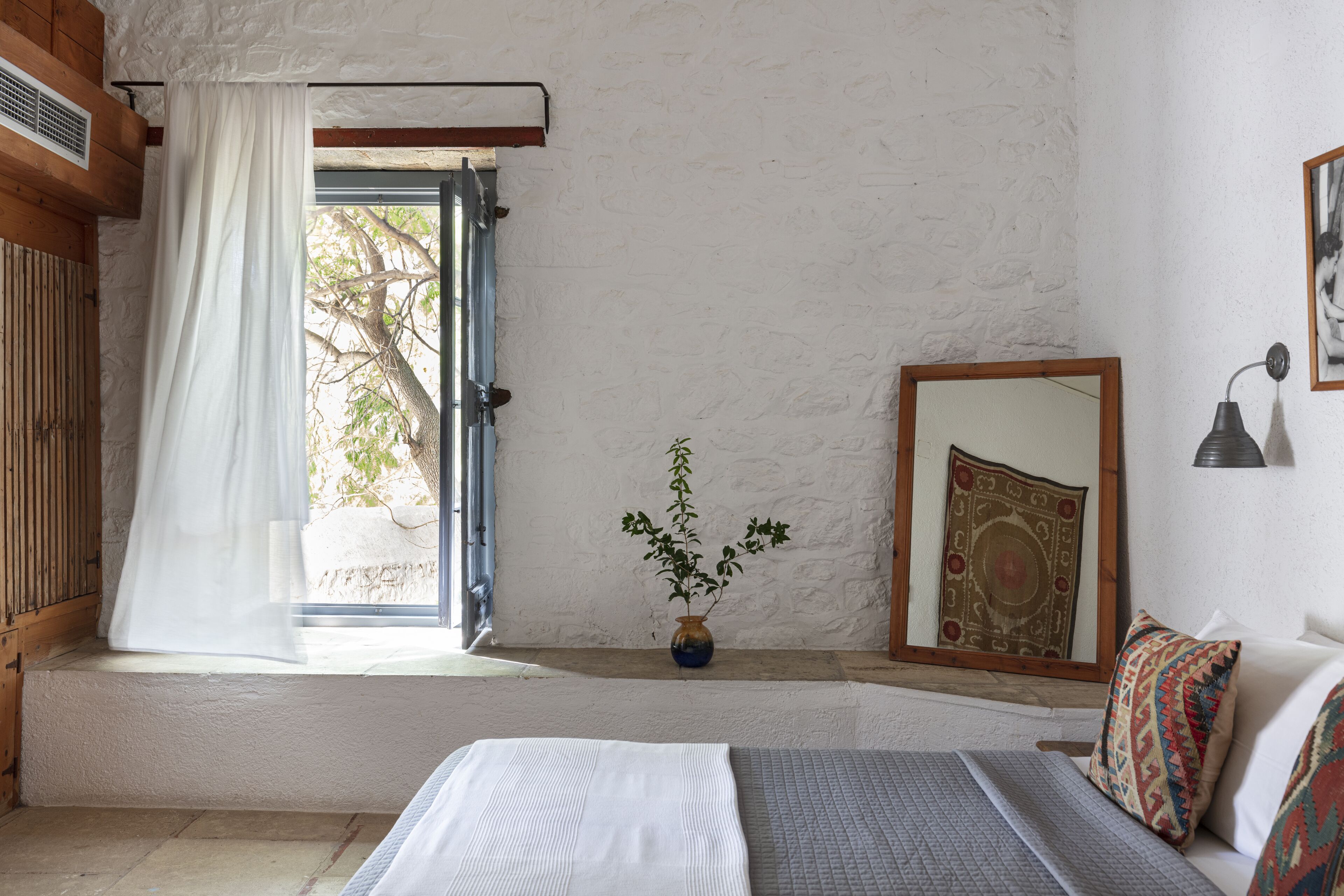 A bedroom with a bed, a window with curtains, a vase with a plant, a mirror, and a wall-mounted lamp.