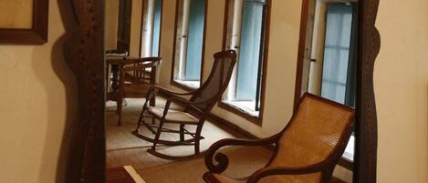 A large mirror with an ornate frame reflecting a room with a wooden ceiling, a chandelier, and a rocking chair.