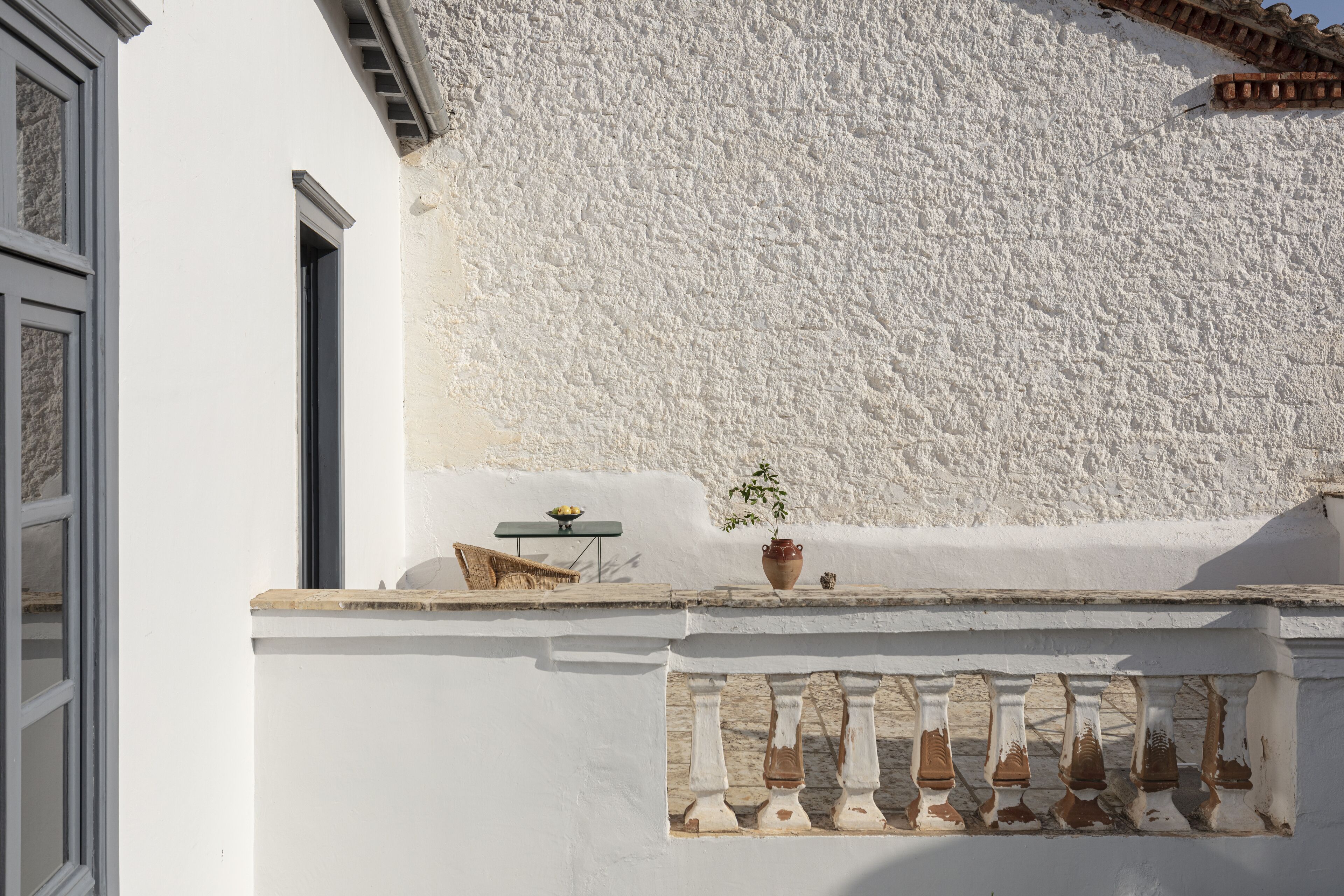 A white building facade with a textured wall, a small table with a bowl and a potted plant, and a balcony railing with a sign.