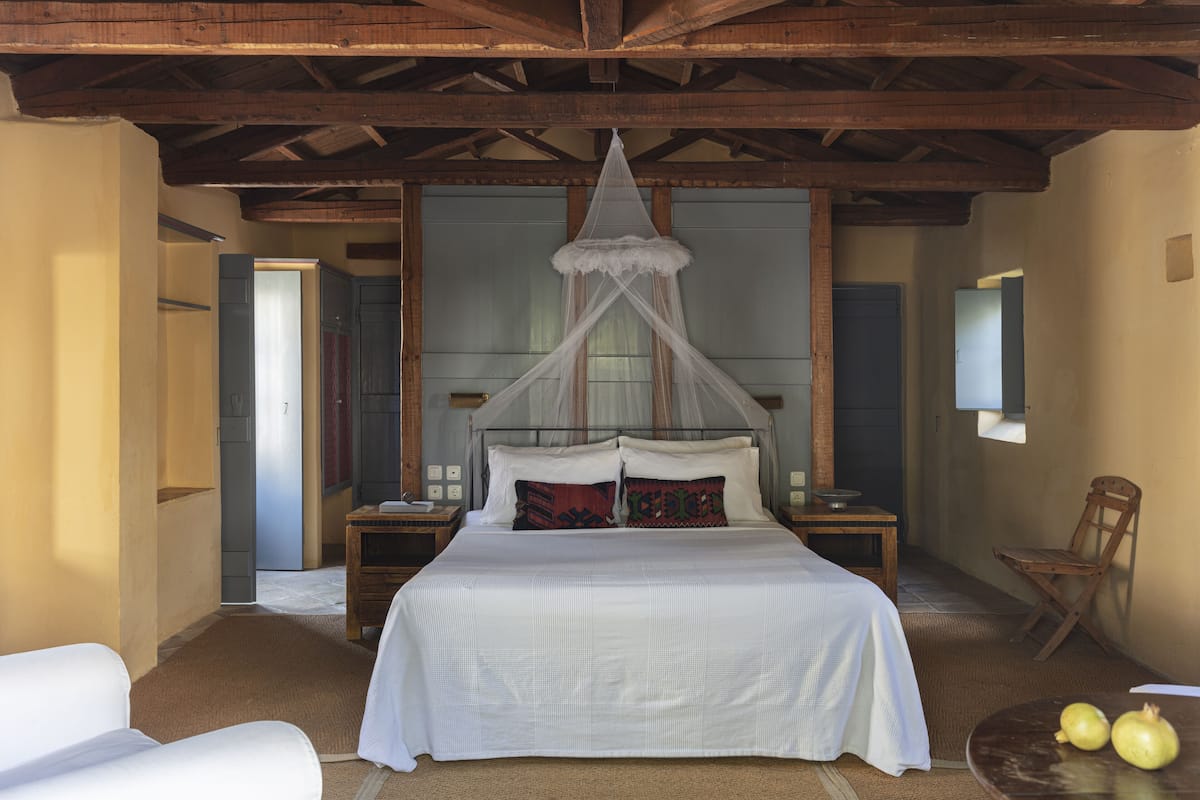 Une chambre à coucher avec un grand lit, une tête de lit en bois et un baldaquin. On y trouve deux tables de chevet avec des lampes, une chaise et une petite table sur laquelle reposent deux grenades.